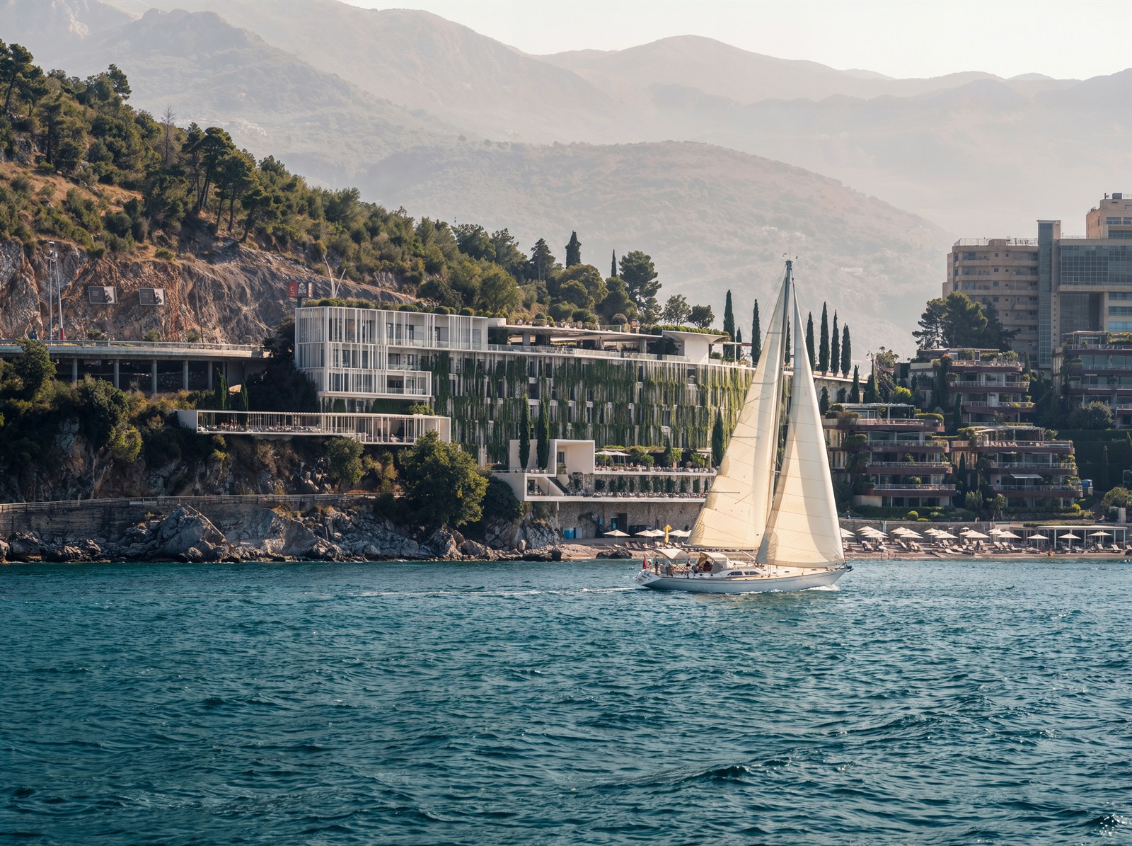 Sailboat passing the green-clad resort under hazy mountain layers